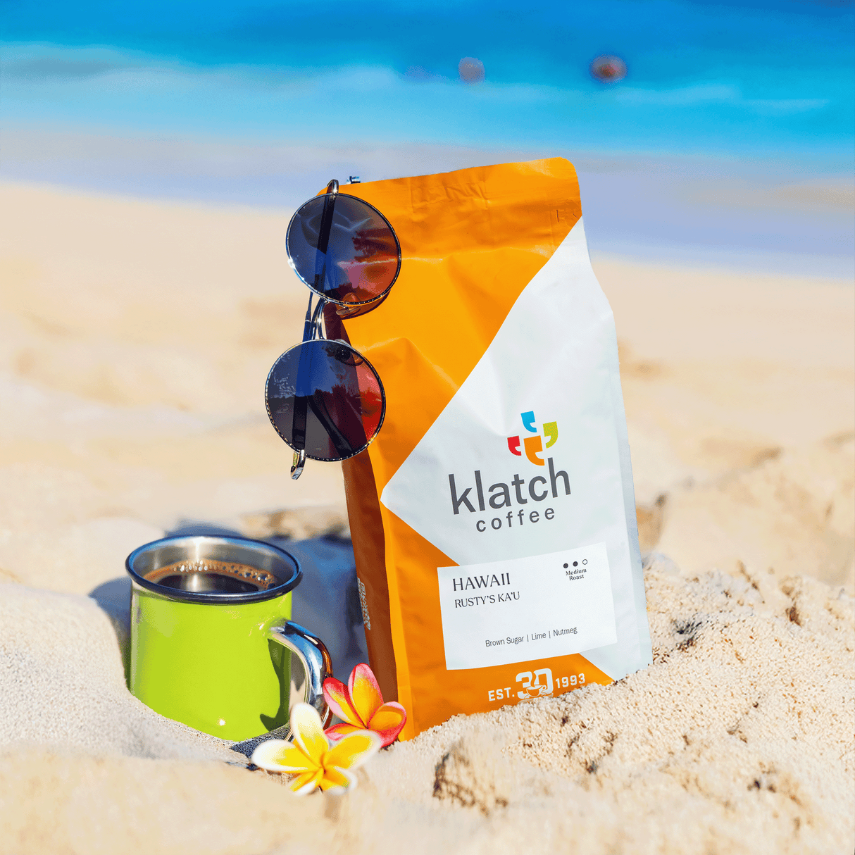 Klatch Coffee bag with sunglasses and a mug on sand with ocean background