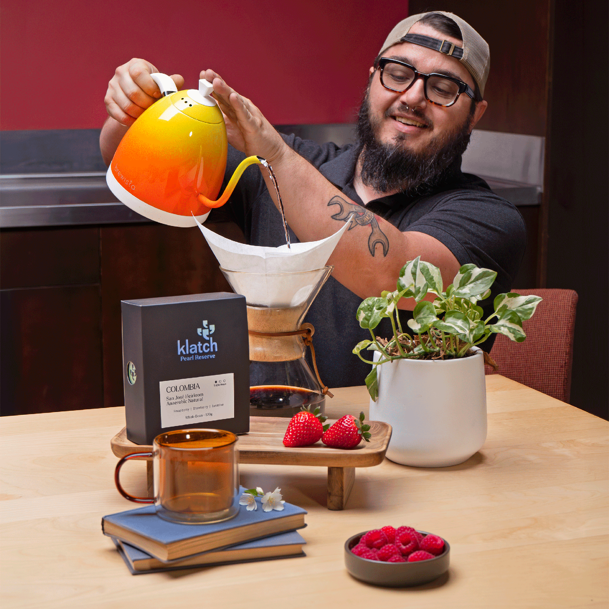 Man making our Colombia Heirloom Anaerobic Natural coffee with a chemex pour over using an orange electric kettle on a table with a plant and berries.