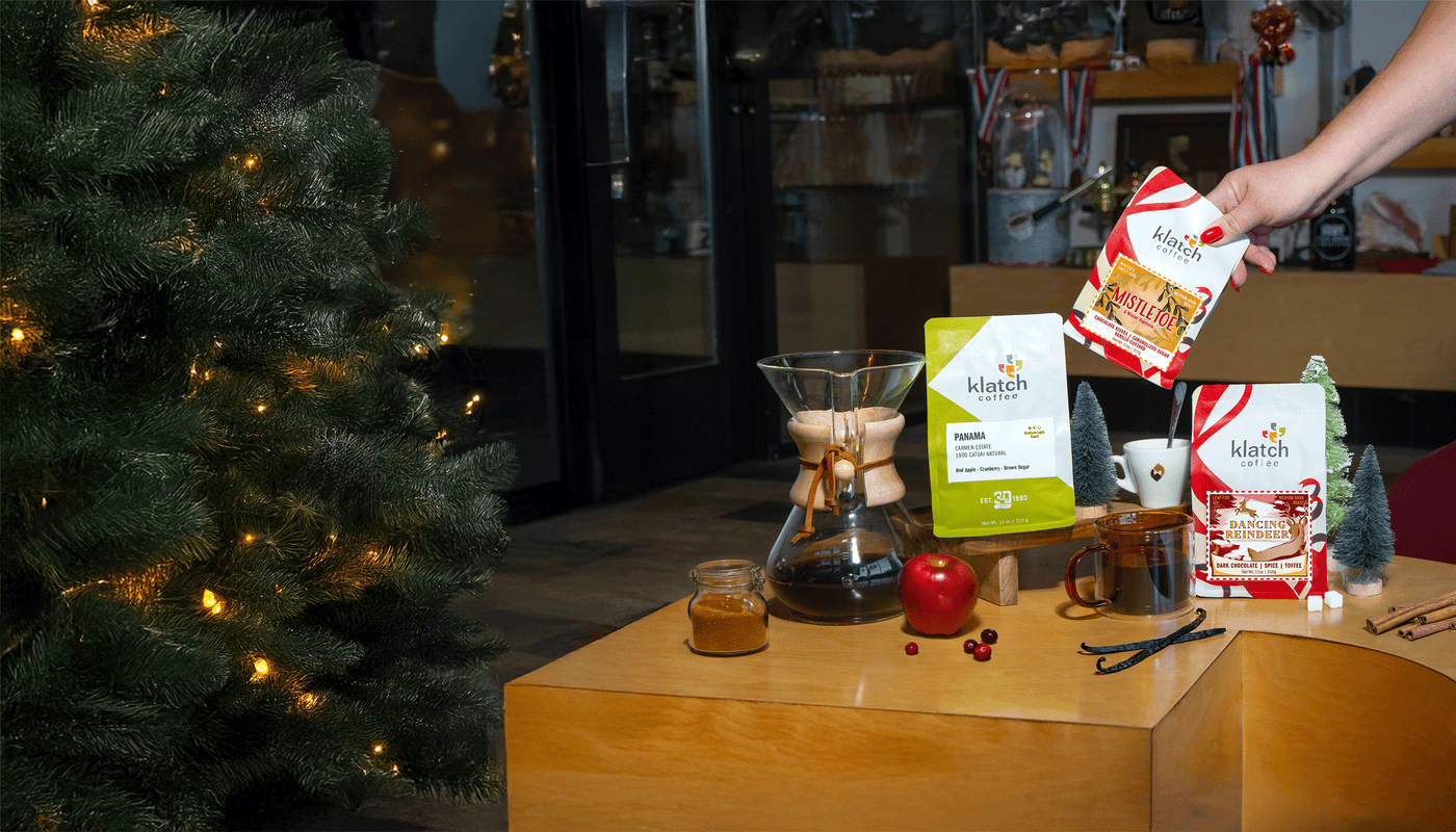 Person holding coffee bag with November Club selections from a table with a Christmas tree in the background.