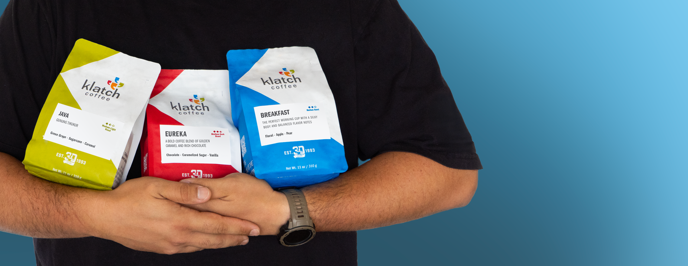 Man in a black shirt holding three coffee bags, of coffee club, Java, Eureka and breakfast standing in front of a blue gradient wall.