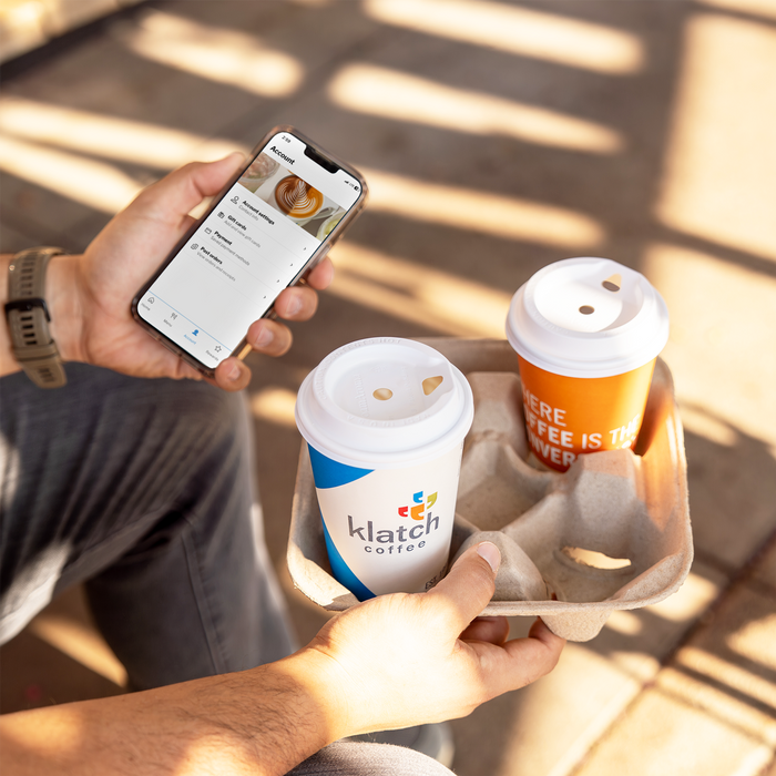 Person holding a phone with the new Klatch Coffee mobile app open in one hand and a drink carrier with two coffees in the other.