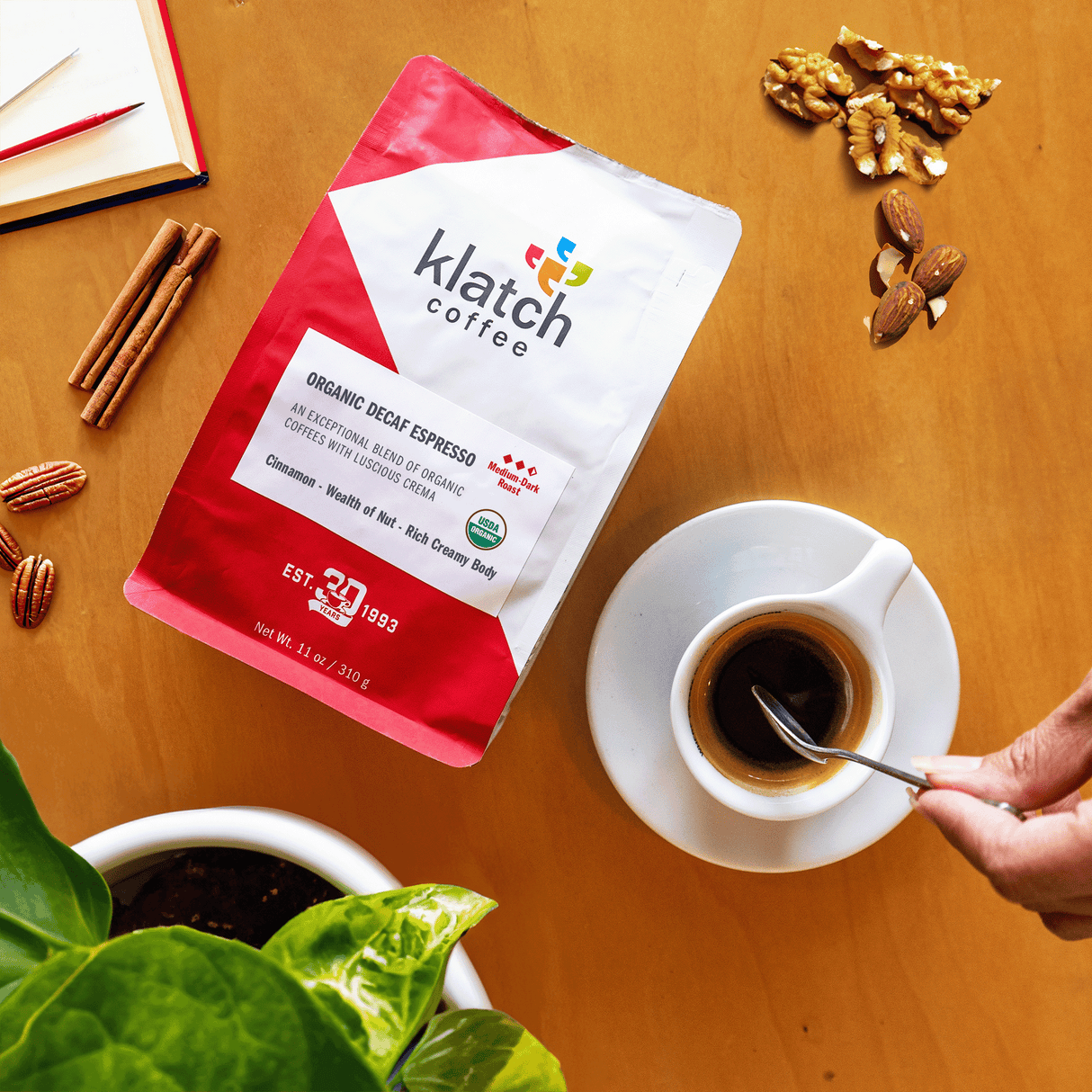 Organic Decaf Espresso bag laying face up on a wooden table qith espresso white ceramic cup being stirred with an espresso spoon by a woman's hand. Cinnamon sticks, walnuts, almonds and pecans lay on the table surrounding coffee bag.