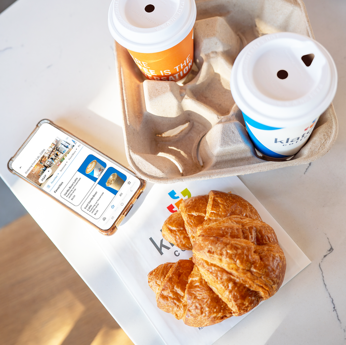 A smartphone displaying the new Klatch Coffee® mobile app rests on a table beside two drinks and a fresh croissant.