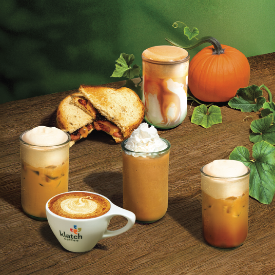 A collection of seasonal coffee beverages and a toasted bacon, cheddar, and apple butter sandwich arranged on a rustic wooden table with a pumpkin and green vines in the background, celebrating the cozy flavors of fall.