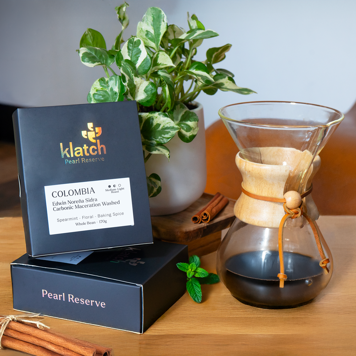 Grey pearl box stacked on another beside a plant on a wooden stool, with a Chemex of coffee, cinnamon sticks, and spearmint leaves nearby.