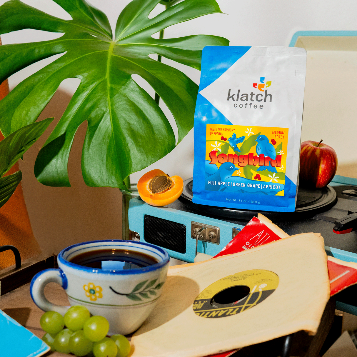 Songbird coffee bag on record player with apple and apricot. A wooden tray with a cup of coffee, grapes with record. A large leave plant in the background.