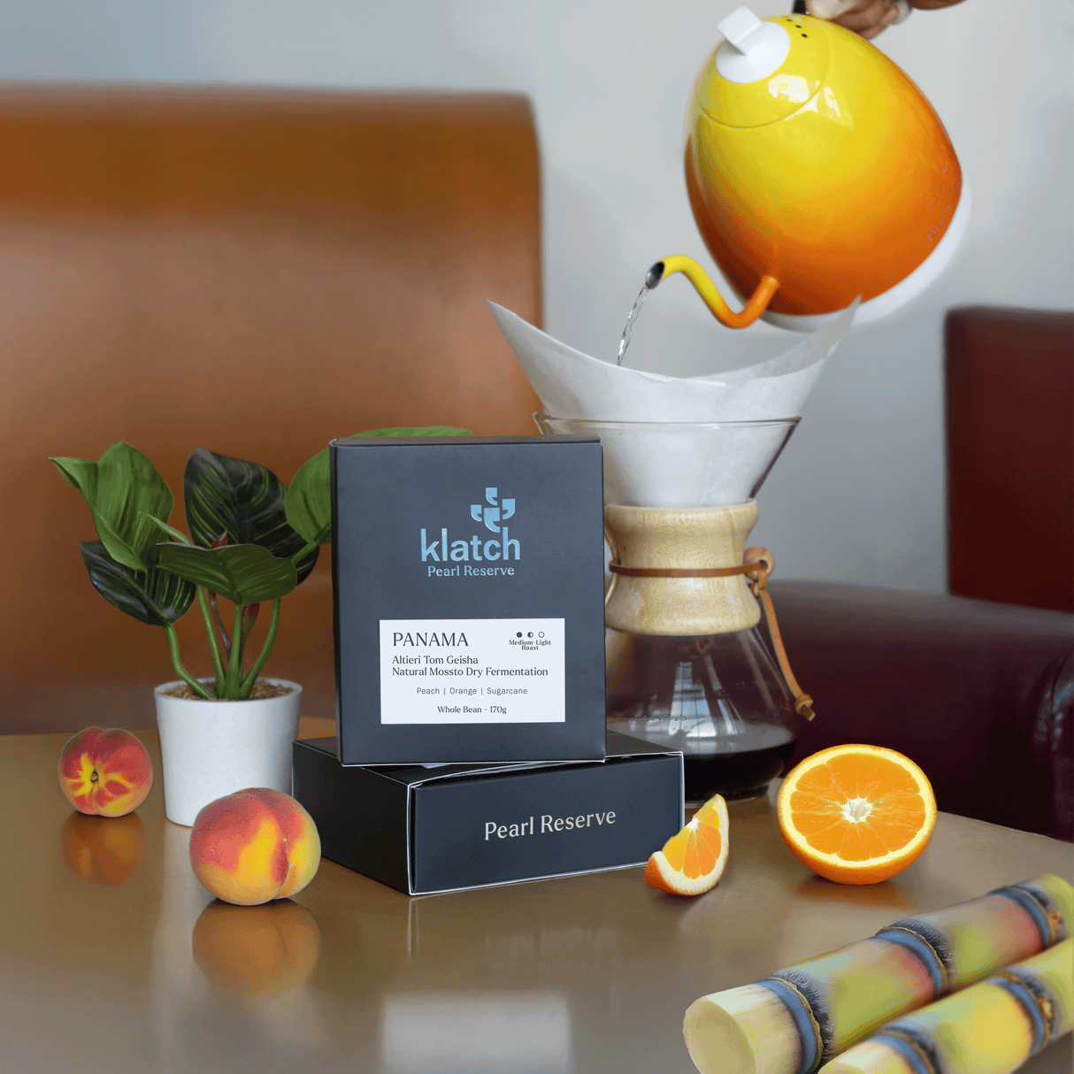Table with a Panama Altieri Tom Geisha box with a orange-yellow kettle pouring hot water into a chemex pour over, peaches, and oranges.