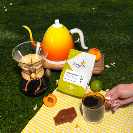 Organic Sumatra coffee bag set on yellow blaet with kettle, chemex and hand holding coffee mug on grass.