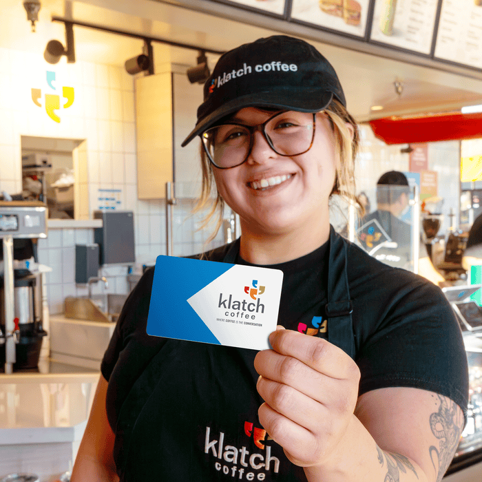 A barista stands in a café holding a gift card that has “Klatch Coffee.” written on it.