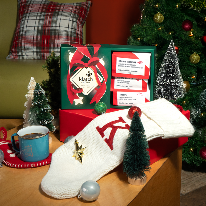 Holiday Coffee Trio on gift box with white stocking with red 'K', tiny christmas trees, ortamintes, teal metal coffee mug with christmas tree in background.