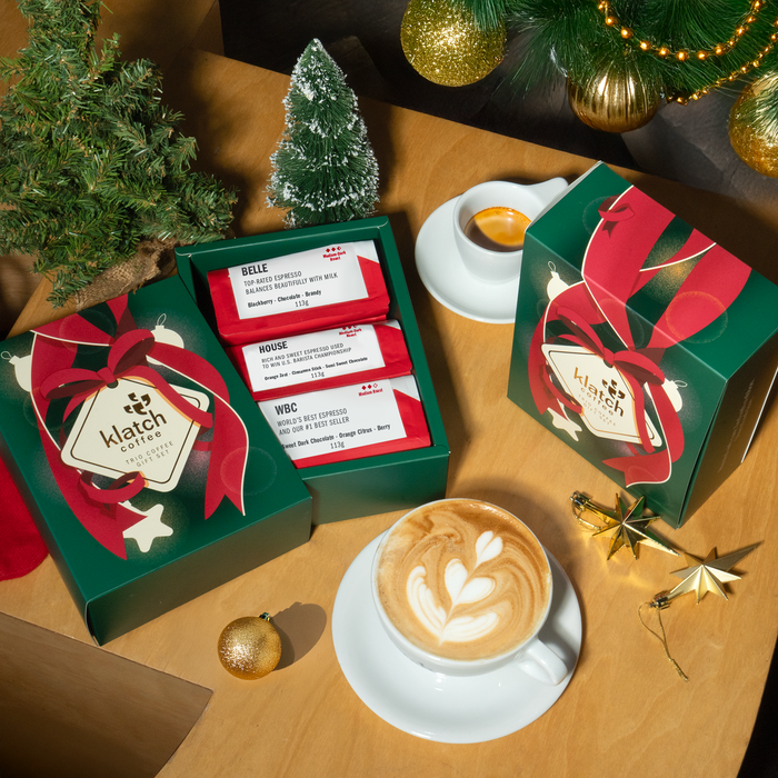 Top view of open and closed green Holiday Trio Coffee boxes, with small pine trees, star ornaments, a latte in a white cup on saucer and an espresso in a white cup on saucer all placed on table.