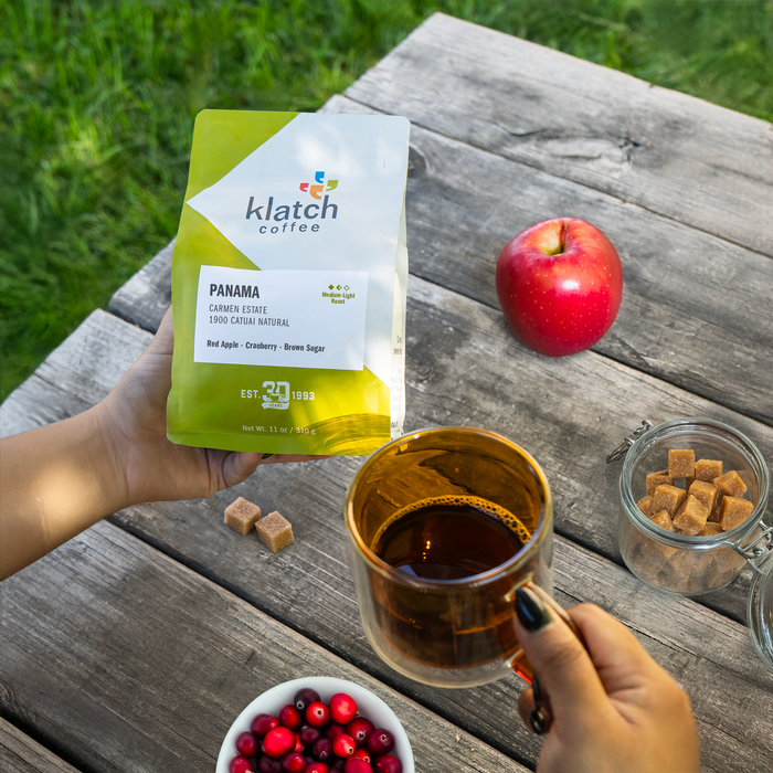 Person holding a green Panama coffee bag in one hand and a coffee cup in the other, with an apple, brown sugar, and cranberries on the table.