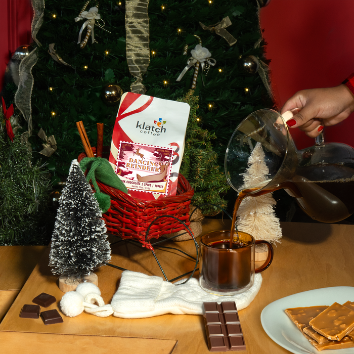 Dancing Reindeer coffee bag in a red sleigh with someone pouring coffee into a mug, surrounded by Christmas decorations and chocolate.