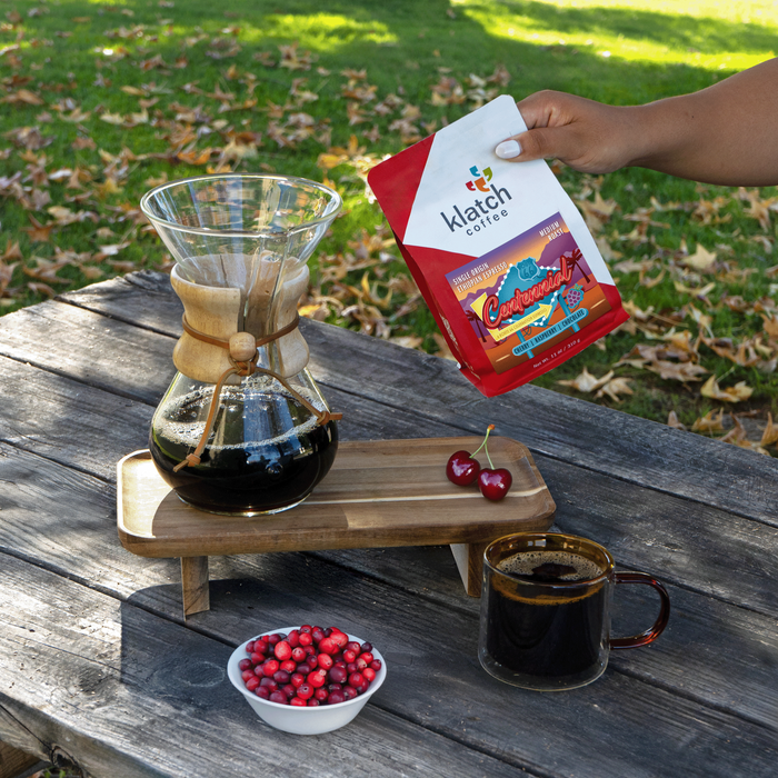 January Club selection of Single Origin Centennial coffee bag held over of picnic table with chemex of coffee, glass mug of coffee with berries on tray stand and bol of cranberries. With grass  in the background.