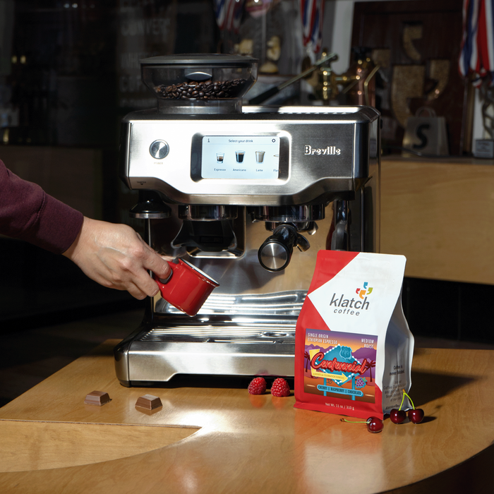 Breville espresso machine with a hand holding a red espresso mug, next to a bag of klatch coffee Centennial on a wooden table.