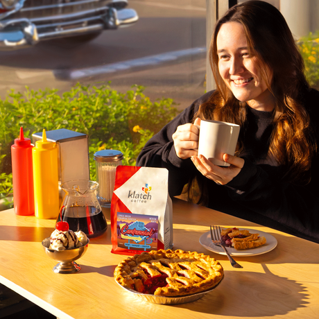 Woman holding a coffee cup with a pie, ice cream, and a Klatch Coffee bag of Centennial on a table in diner.