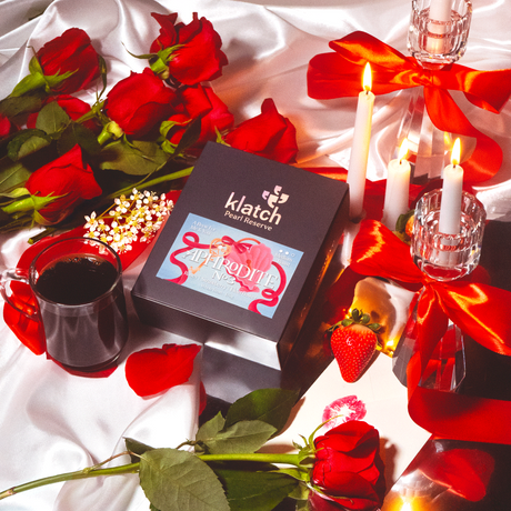 Valentine's Day-themed arrangement with red roses, candles, and a box of Klatch Coffee Aphrodite on a white fabric background.