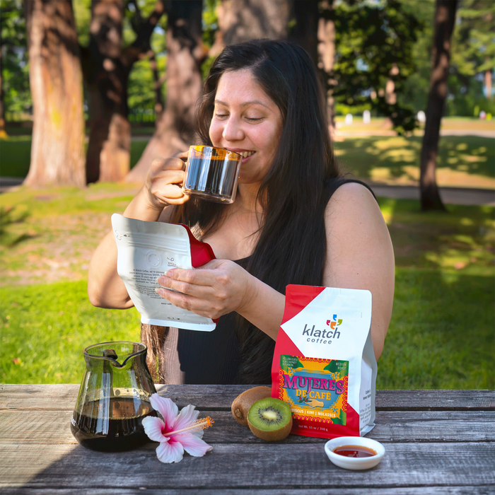 Woman enjoying a cup of coffee outdoors with a Mujeres De Cafe bag and other items on a table.