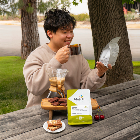 Person enjoying coffee outdoors with a Klatch coffee package on a wooden table.