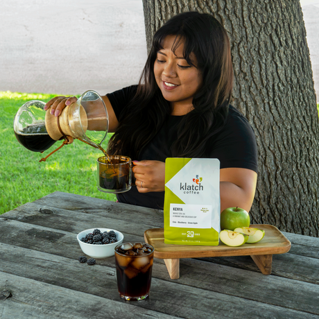 Woman pouring coffee from Chemex into glass mug. A bag of Kenya Wang'era AA set on tray with green apple on a picnic table; with blackberries in a white bowl next to a cola in a glass cup.