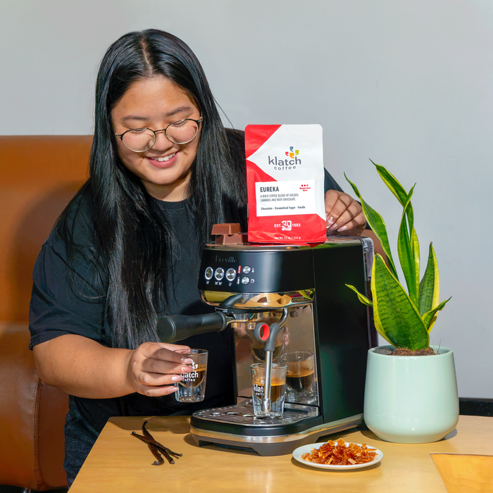 Woman preparing espresso with a Breville espresso machine using Klatch Coffee Eureka blend, with branded espresso glasses and coffee ingredients nearby.