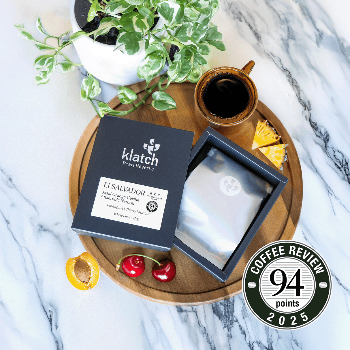 Klatch coffee El Salvador Jasal Orange Geisha - 94pts Coffee review badge package on a wooden tray with a cup of coffee, plant, and fruit on a marble background
