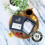 Klatch coffee El Salvador Jasal Orange Geisha - 94pts Coffee review badge package on a wooden tray with a cup of coffee, plant, and fruit on a marble background