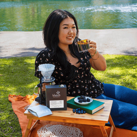 Woman sitting outdoors by a lake, holding a coffee cup with a coffee-making setup on a small table.