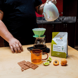 Person pouring coffee into a green pour-over coffee maker with a bag of klatch coffee and fruits on a wooden table.