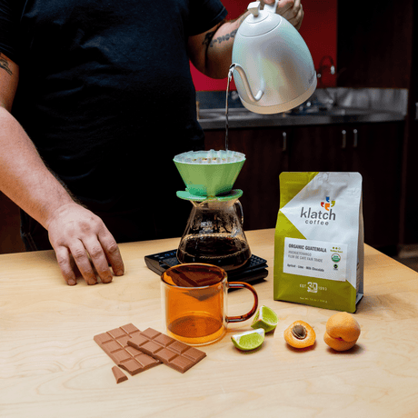 Person pouring coffee into a green pour-over coffee maker with a bag of klatch coffee and fruits on a wooden table.