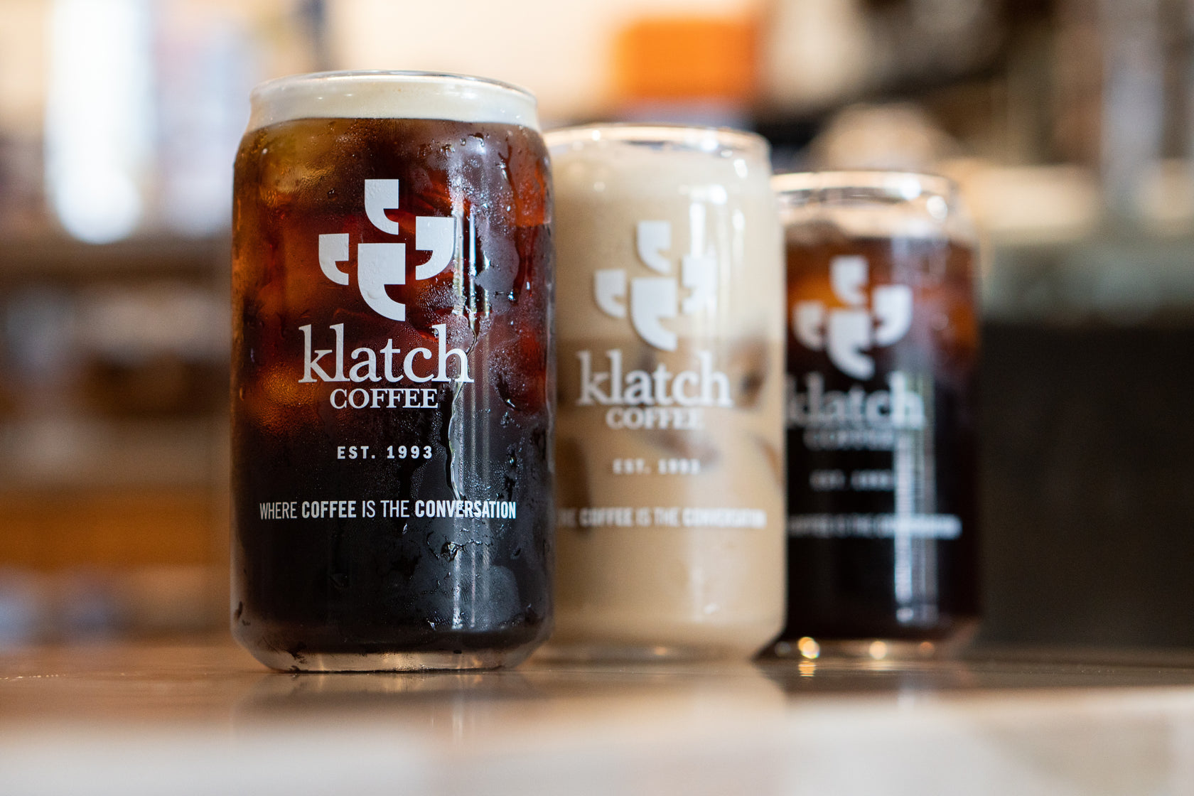 Three iced drinks in clear glass cups with the Klatch Coffee logo on each glass, arranged on a café counter.