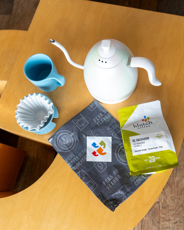 Coffee bag on wooden table with klatch delivery bag. White kettle, blue mug and blue brewing device on the side.