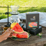 A hand holds a slice of watermelon beside a black Klatch Panama Elida Aguacate Geisha Natural box and glass pour-over coffee equipment on a wooden table, with a blurred green lawn in the background.