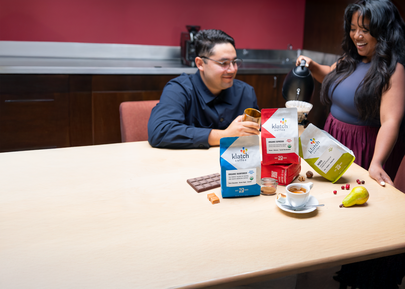 Two people sitting at a table with Klatch coffee packages of Aug club coffees and a cup of coffee.