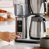 Stainless steel coffee maker on a kitchen counter with a hand adjusting settings.