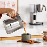 Person pouring coffee from a Bialetti coffee maker into a cup on a kitchen counter.