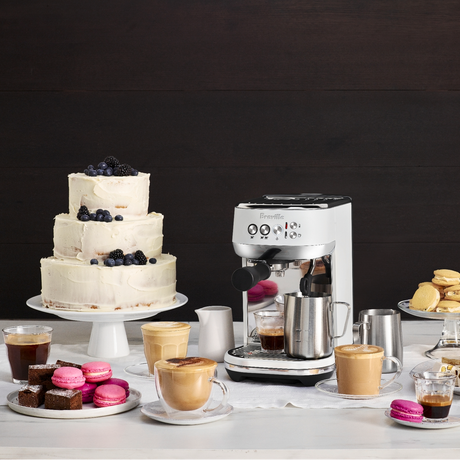 Breville Bambino Plus Espresso Machine with a cake, cookies, and coffee cups on a table against a dark background