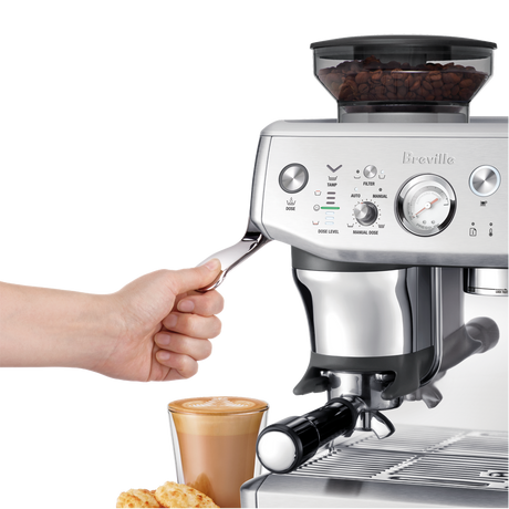 Breville Barista Express Impress Espresso Machine with a hand holding a scoop of coffee beans and a cup of coffee in the background.