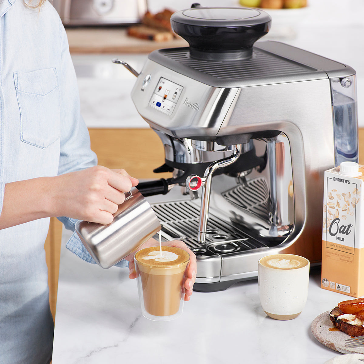 Person pouring steamed milk from pitcher in to double walled glass. The Breville Barista Impress Touch Espresso Machine is behind pour.