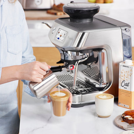 Person pouring steamed milk from pitcher in to double walled glass. The Breville Barista Impress Touch Espresso Machine is behind pour.