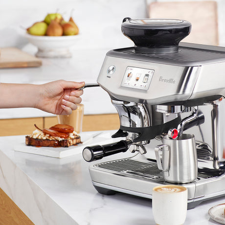 Breville Impress Touch Espresso Machine with a digital display. A ceramic mug with a latte, steam wand in the pitcher steaming milk on a white counter.