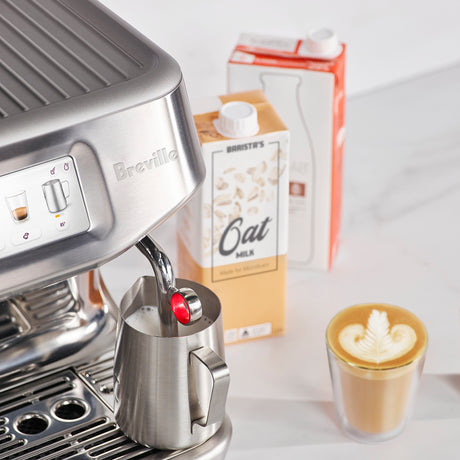 Breville Impress Touch Espresso Machine with a digital display. A glass of latte, pitcher steaming milk and carton of Oat milk on a white counter.