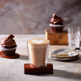 Glass of cappuccino with a heart foam on a wooden coaster, surrounded by cupcakes and a glass on a rustic surface.