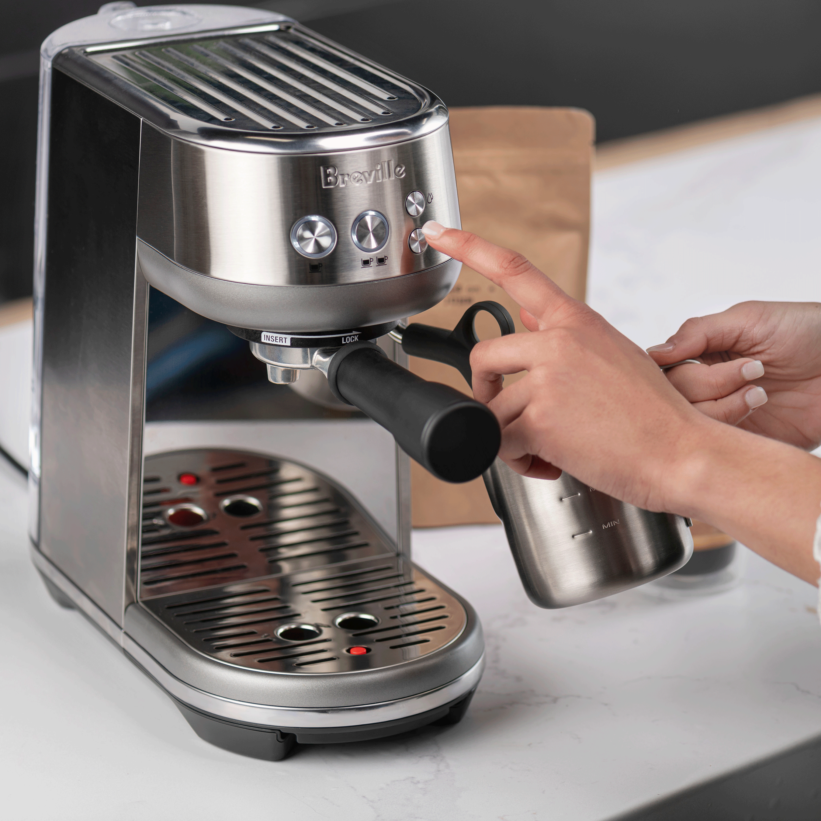 Hands operating a Breville Bambino Plus espresso machine while holding a stainless steel milk pitcher.