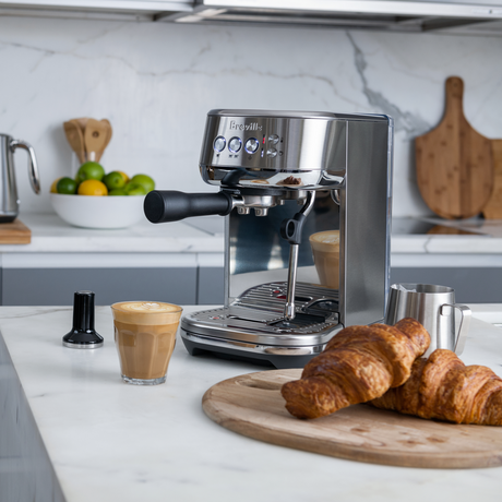 Breville Bambino Plus Espresso Machine on a kitchen counter with croissants and coffee.