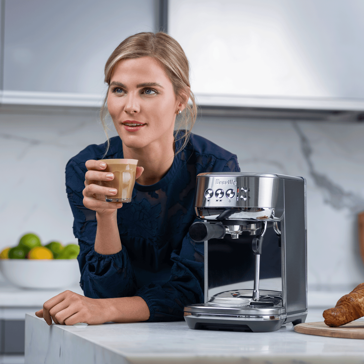 Woman holding a latte beside a Breville Bambino Plus espresso machine on a kitchen counter.