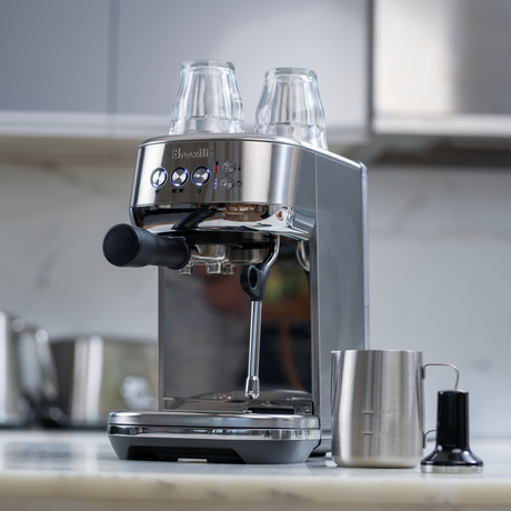 Breville Bambino Plus Espresso Machine with two classes set on top for storage with the pitch and tamper on a kitchen counter beside it with blurred background