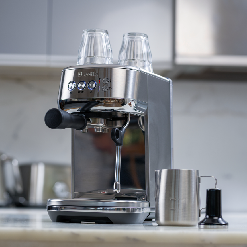 Breville Bambino Plus espresso machine with portafilter, steam wand, stainless steel milk pitcher, and tamper on a kitchen counter.