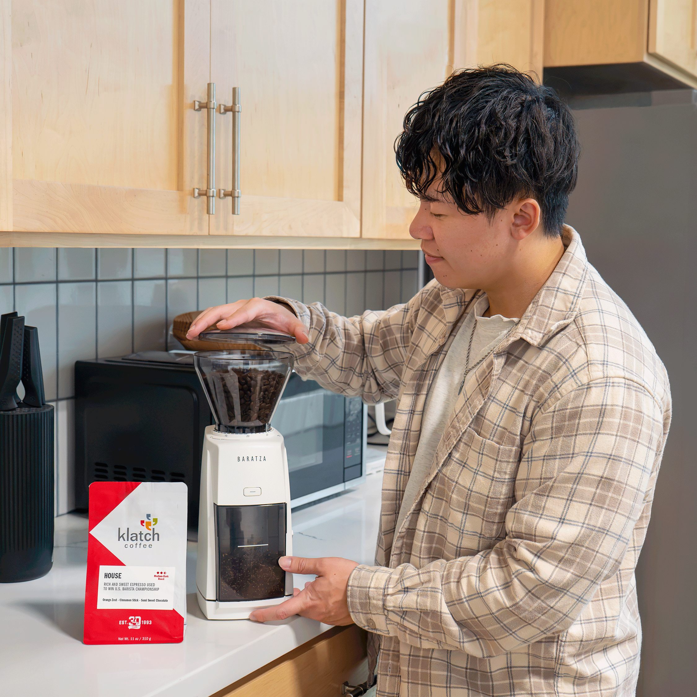 Man using a Baratza coffee grinder alongside a bag of Klatch Coffee House Espresso in a home kitchen.