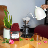 Red velvet drawstring coffee bag, with pour over, potted plant, pink rose, white cherries and spearmint on a wooden coffee table.